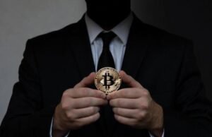 A shadowy anonymous figure in a black suit and tie, face hidden in darkness, holds a golden Bitcoin coin prominently, evoking the mystery of Satoshi Nakamoto — the unknown creator at the heart of crypto history.